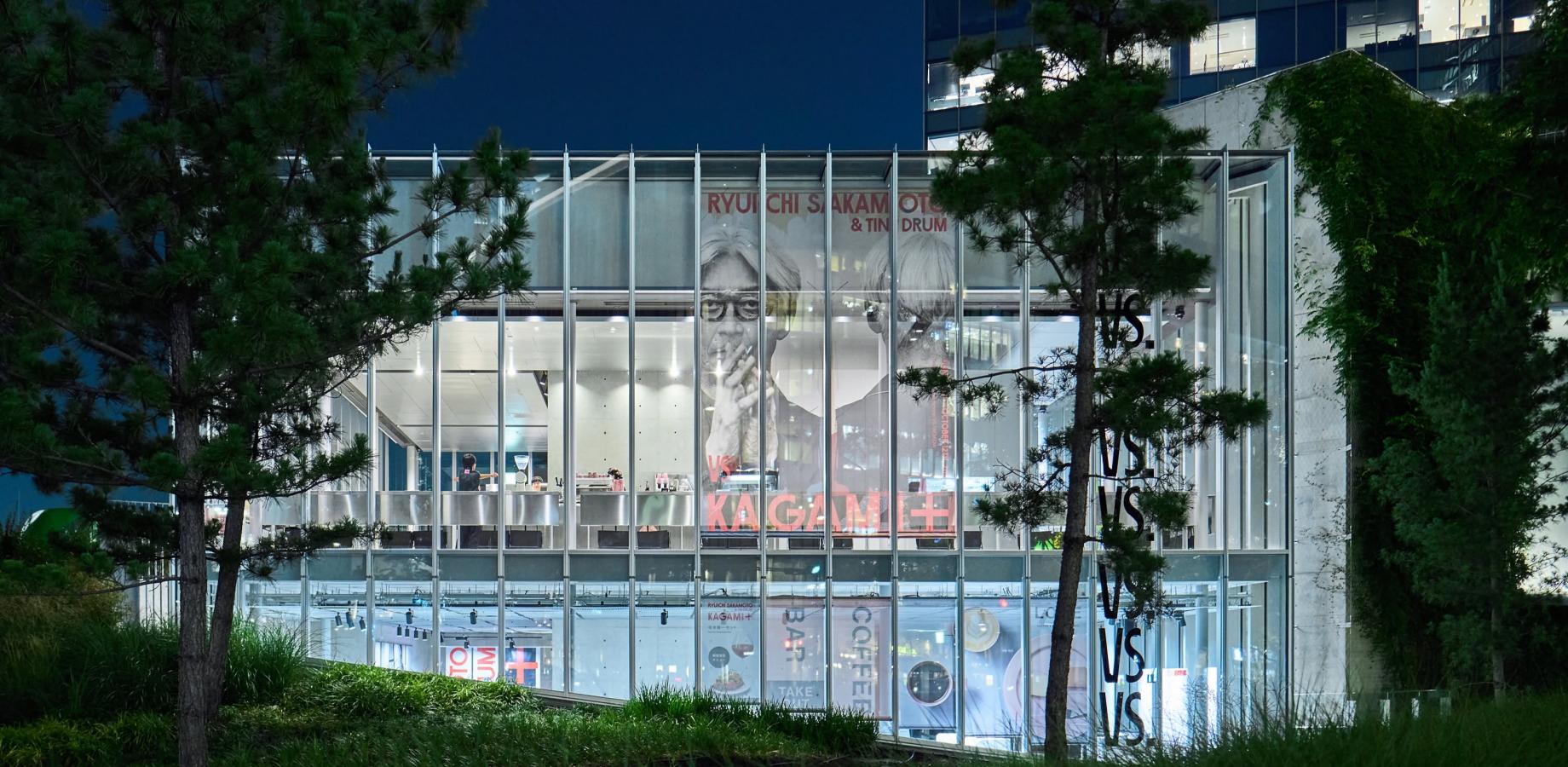 Glass-fronted KAGAMI exhibition building in Osaka at dusk, surrounded by trees