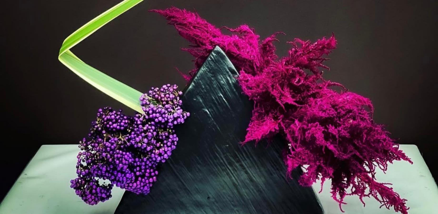 Ikebana arrangement of purple berries and magenta plumes with a green leaf on a dark triangular base
